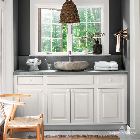 A white bathroom vanity with stone vessel sink underneath a charcoal gray wall with window.