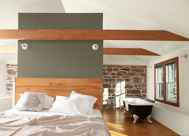 White loft bedroom, sage green accent wall, wooden beams, stone wall, freestanding bathtub.