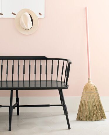 Pared rosa con escoba, banco de silla negra y sombrero sobre perchero blanco.