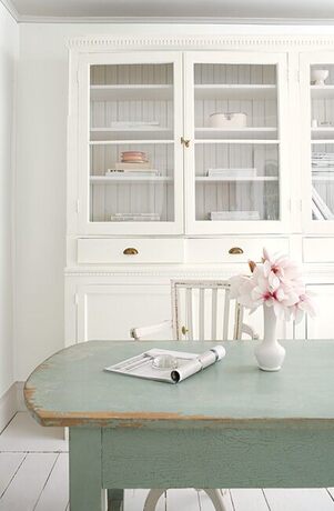 Shabby chic dining: white walls, cabinet, chair; blue table, flowers; light gray wood floor.