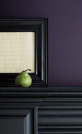 A rich, purple-painted wall with a black framed art piece and a green pear atop a sleek black mantel