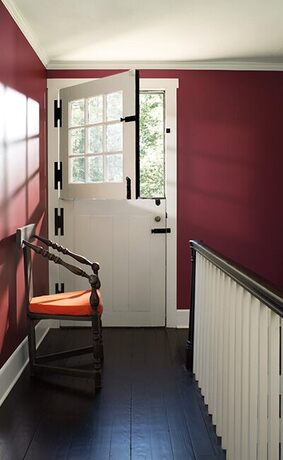 White dutch door, sunlit burgundy hallway, orange cushioned chair, dark wood floor.
