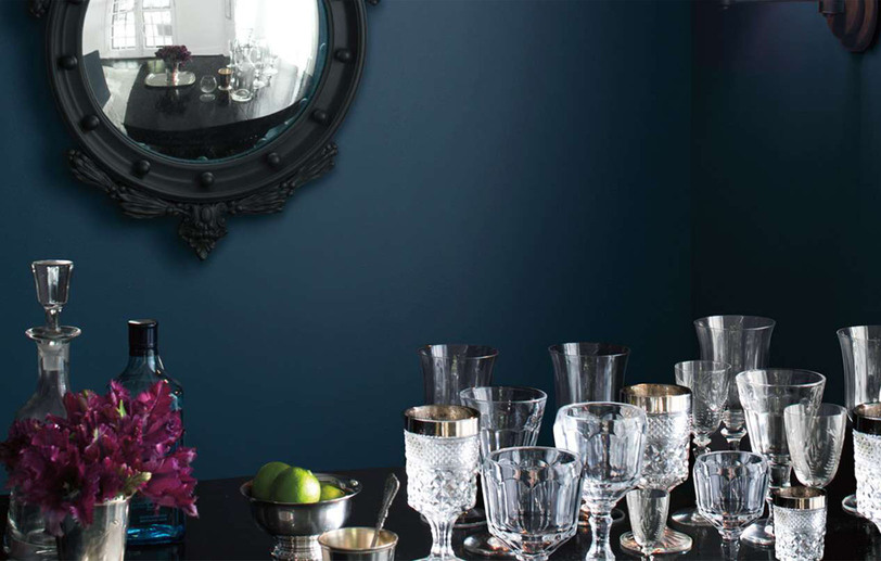A dark blue-painted room with black framed mirror and crystal glasses on a table.