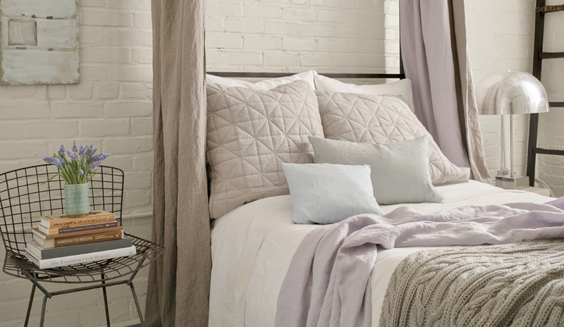 Bedroom with a white washed brick wall behind a tan canopy bed with lilac pillows and throw blanket.