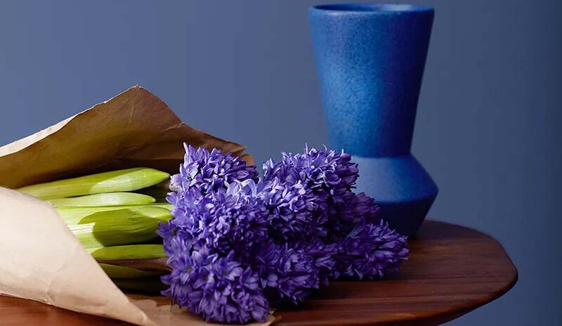 Deep blue wall with decorative trim, small table, cobalt vase, purple bouquet in brown paper.