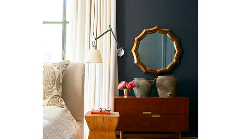 Un dormitorio pintado de negro con cama crema, mesas decorativas de madera con macetas y flores