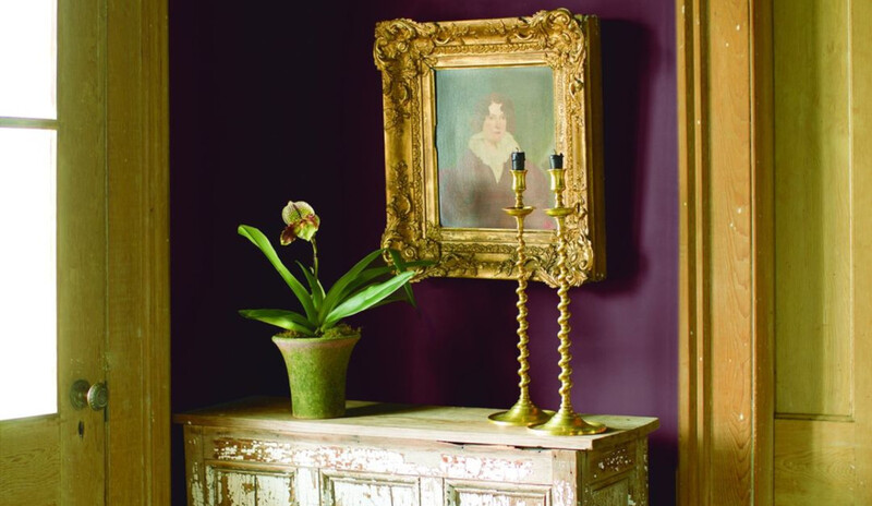 Entrada luminosa con velas, flor en maceta sobre mesa desgastada y pared de color ciruela.