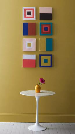 Pequeña mesa blanca frente a una variedad de cuadros cuadrados en una pared de Savannah Green.