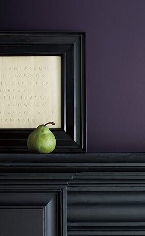 A rich, purple-painted wall with a black framed art piece and a green pear atop a sleek black mantel