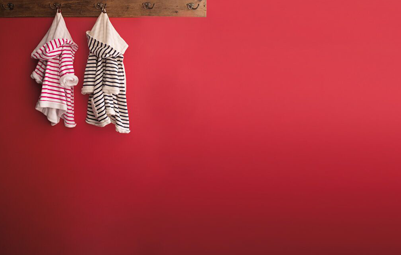 Pared roja con un perchero de madera, dos batas de rayas para niños, una en rosa y otra en negro.