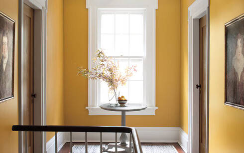 Pasillo pintado de amarillo con mesa de madera y flores, dos puertas, una ventana y marcos pintados 