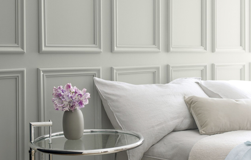 Gray wall with gray bedding and a glass side table featuring a small vase of purple flowers.