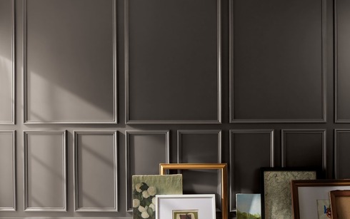 Artwork and frames arranged on a floor against a deep chocolate brown-painted wall with decorative